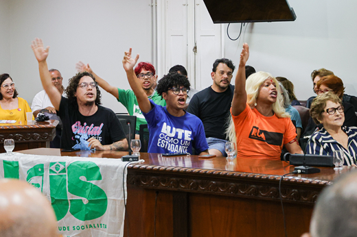 Imagem: Em primeiro plano três estudantes erguem o braço e gritam palavras de ordem na sala do CONSUNI