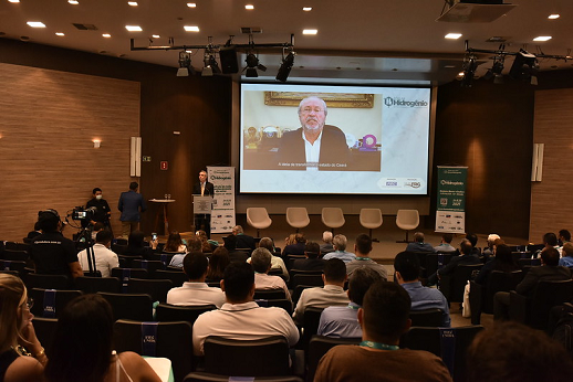 O reitor da UFC, Prof. Cândido Albuquerque, participou da abertura do evento por meio de vídeo reproduzido no auditório da FIEC (Foto: Viktor Braga/UFC) Imagem mostra telão onde aparece o reitor da UFC em auditório amplo com pessoas sentadas de costas para o fotógrafo e acompanhando o discurso