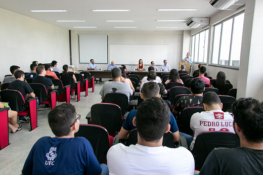 Durante cerimônia, o reitor da UFC, Prof. Cândido Albuquerque, fez uma prestação de contas da gestão (Foto: Viktor Braga/UFC) Imagem: Pessoas sentadas em cadeiras no auditório, com uma mesa de autoridades adiante
