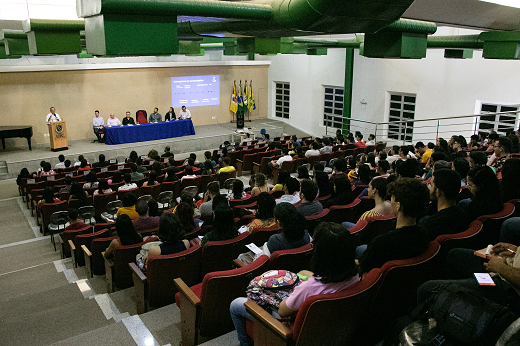 A cerimônia de recepção dos novos alunos ocorreu na unidade Mucambinho, do Campus de Sobral (Foto: Ribamar Neto/UFC) Imagem: Alunos sentados em um auditório; à frente, na parte elevada, está a mesa do evento composta por professores da UFC; o reitor está de pé, falando ao microfone (Foto: Ribamar Neto/UFC)