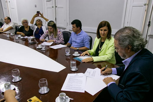 Momento da assinatura do protocolo de intenções pela presidente da APDMCE, Tamara Bezerra (quarta da esquerda para a direita) e pelo reitor Cândido Albuquerque (Foto: Viktor Braga/UFC) Imagem: Várias pessoas sentadas em torno de uma mesa, no momento da assinatura do protocolo de intenções (Foto: Viktor Braga/UFC)