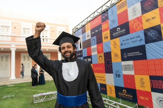 Mark Allison realizou o sonho de voltar a morar no Ceará e estudar na UFC (Foto: Viktor Braga/UFC) Fotografia do concludente Mark Allyson comemorando colação em frente a painel da UFC