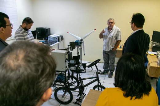 Os visitantes tiveram a oportunidade de conhecer um protótipo de um triciclo assistivo para crianças com hemiplegia (Foto: Ribamar Neto/UFC Informa) Imagem: Um grupo de pesquisadores em pé, sendo apresentados a um protótipo de um triciclo assistivo para crianças com hemiplegia