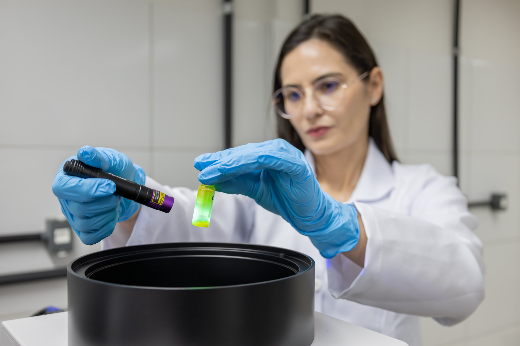 Imagem: foto mostra uma mulher de jaleco branco e óculos de grau segurando um pequeno vidro com substância verde e em outra mão, um dispositivo cilíndrico