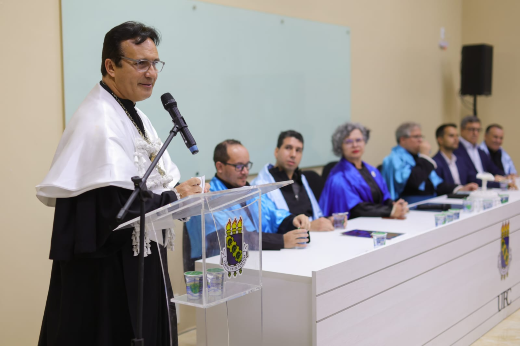 Imagem: foto do reitor vestindo veste talar com capa branca e falando ao microfone no púlpito do auditório