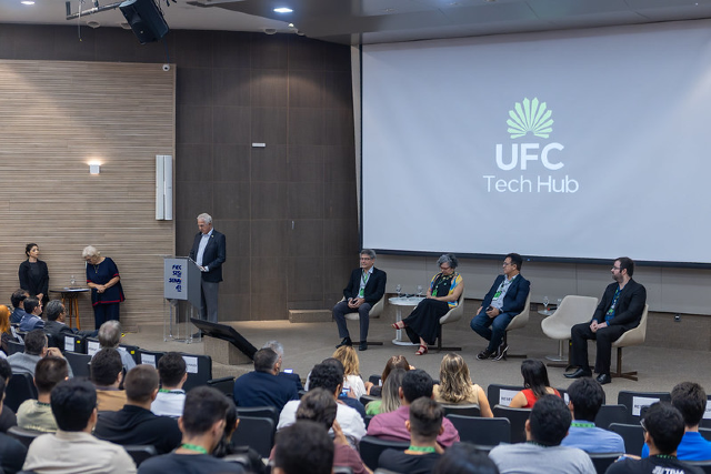 O novo hub é fruto de uma parceria estratégica entre a UFC e a empresa Neo Ventures (Foto: Guilherme Silva/UFC Informa) Imagem: Foto mostra a vista do palco de um grande auditório onde há pessoas sentadas nas cadeiras olhando para o palco, que tem representantes da UFC sentados e um telão atrás deles onde se pode ler Tech HUb UFC