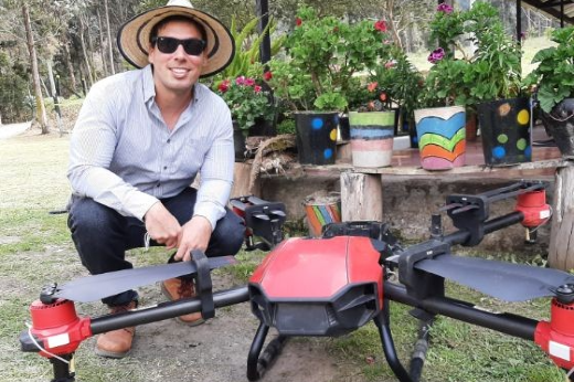Imagem: foto do doutorando da UFC, que veste blusa de mangas compridas na cor branca, chapéu de palha e tênis escuro. está agachado e há um drone grande em frente a ele