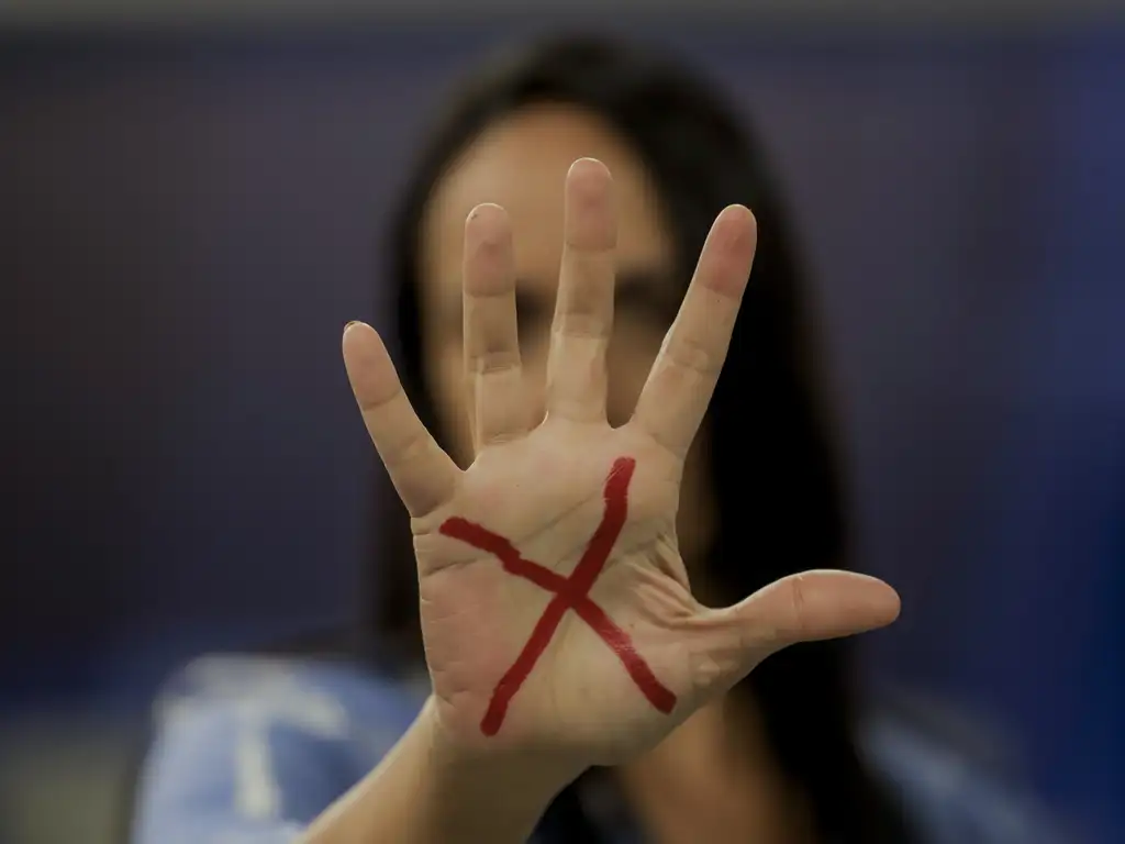 Imagem: Em primeiro plano, mão de mulher mostrando marcação em x, com rosto desfocado