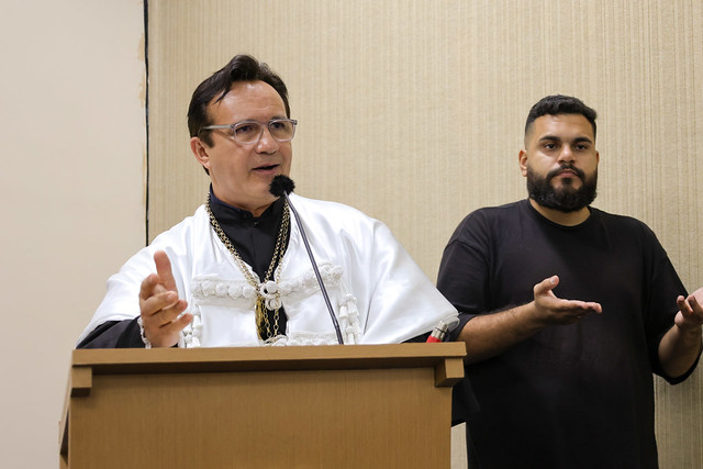 Imagem: Um homem veste beca branca e fala em um púlpito, gesticulando com as mãos. Ao lado dele, um intérprete de Libras, de camiseta preta, traduz o discurso.