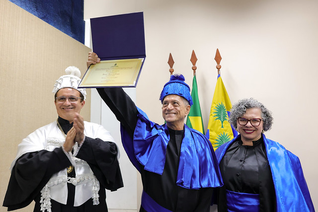 Imagem: Três pessoas posam em uma cerimônia. O homem ao centro, vestindo beca e barrete azul, ergue um diploma com orgulho. À esquerda, um homem de beca branca aplaude, e à direita, uma mulher de beca azul sorri. Ao fundo, bandeiras do Brasil e de um estado.