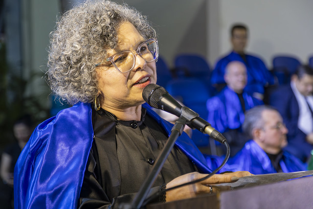 Ao se dirigir aos formandos, Diana Azevedo, vice-reitora da UFC, destacou o papel das famílias e das pessoas próximas na travessia universitária (Foto: Guilherme Silva/UFC) Imagem: uma mulher de cabelos cacheados e grisalhos, vestida de veste talar, fala em um púlpito
