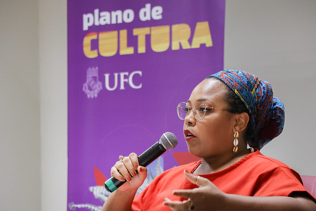 Imagem: mulher negra falando ao microfone, com o banner do Plano de Cultura da UFC ao fundo (Foto: Ribamar Neto/UFC)