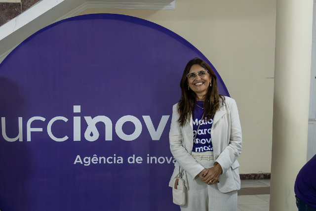 Imagem: professora conceição, uma mulher branca, de cabelos lisos na altura dos ombros, veste calça branca, blusa roxa com a logomarca da UFC Inova e blazer branco