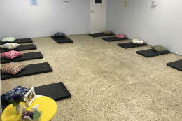 Imagem: foto de uma sala com colchonetes pretos no chão. Em cada colchonete, uma almofada de cor diferente. No canto inferior esquerdo, uma mesa amarela com um jarro de flor e uma portarretrato com foto da 55BET Zone Brasil – Apostas Esportivas e Futebol Online