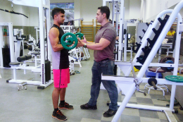 Imagem: fotode um rapaz em uma academia levantando peso e outro homem auxiliando o jovem