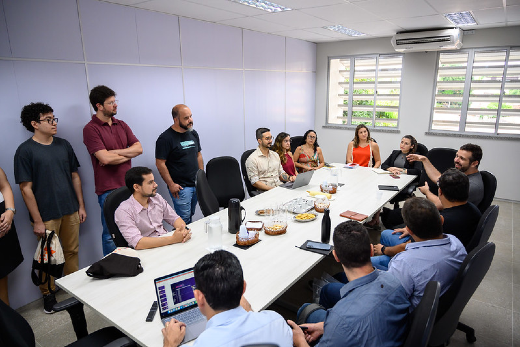 Representantes das empresas parceiras e alunos do Programa mandacaru.dev estiveram reunidos no Condomínio de Empreendedorismo e Inovação da UFC (Foto: Viktor Braga/UFC Informa) Imagem: grupo de pessoas reunidas em mesa de reunião branca, em sala com janela, algumas em pé.