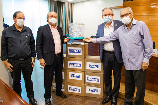 A FIEC anunciou a entrega de 20 unidades do capacete Elmo para o Hospital Universitário Walter Cantídio (HUWC), do Complexo Hospitalar da UFC/EBSERH (Foto: Ribamar Neto/UFC) Imagem: foto de três homens segurando um capacete de respiração assistida