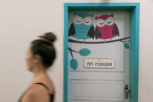 Os PETs contribuem para a formação dos estudantes e ajudam no combate à evasão escolar nas instituições de ensino superior (Foto: Viktor Braga/UFC) Imagem: Mulher de pele branca e cabelos pretos em frente à porta do PET Pedagogia