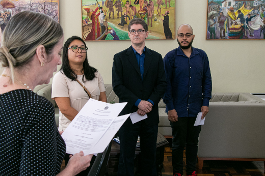 Os novos servidores prestaram o compromisso de cumprir fielmente com os deveres e atribuições do cargo, observando o Código de Ética Profissional dos Servidores da UFC (Foto: Ribamar Neto/UFC) Imagem: homens e mulher em pé ouvem cerimonialista ler termo de posse