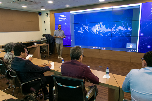 Na apresentação, os gestores da UFC conheceram as ações do Observatório da Indústria e como seu leque de ferramentas de inteligência de dados pode ajudar na elaboração de políticas públicas e compreensão das tendências de mercado. (Foto: Ribamar Neto/ UFC Informa) Imagem: De pé, à frente, o gerente do Observatório da Indústria, Guilherme Muchale, apresenta à comitiva da UFC (sentados em uma bancada). Ele está à frente de um telão azul. (Foto: Ribamar Neto/ UFC Informa)