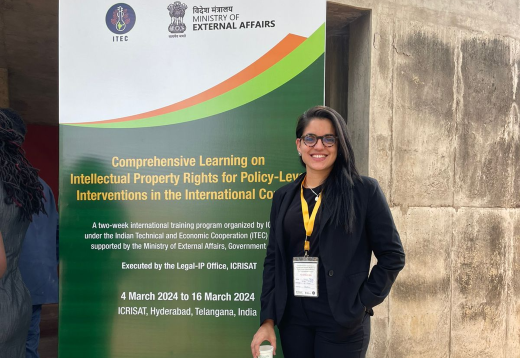 Ana Carolina Matos, servidora da PROINTER, participa de curso sobre direitos de propriedade intelectual na cidade de Telangana, na Índia (Imagem: Arquivo Pessoal) Imagem: Mulher jovem de cabelos pretos e roupa preta em frente a banner de evento