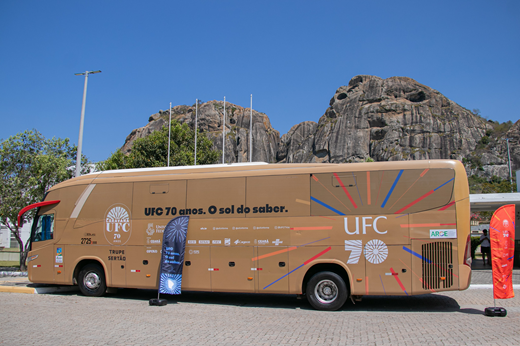 Primeira parada da trupe da Caravana UFC 70 Anos que percorre o Interior foi o município de Quixadá; Campus da UFC preparou programação especial. (Foto: Ribamar Neto/ UFC Informa) Imagem: Vista lateral do ônibus da Caravana UFC 70 Anos (na cor dourado) que está percorrendo os sertões. Ao fundo, os monólitos de Quixadá