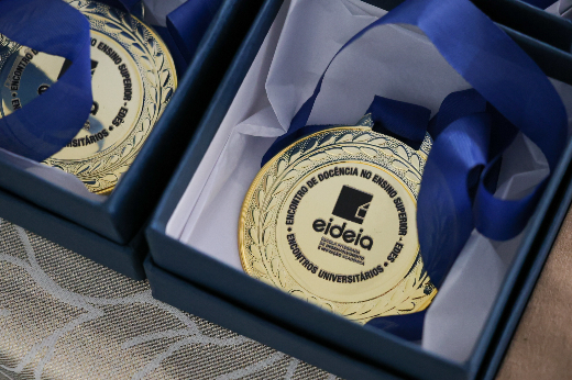 Além da medalha, os oito docentes homenageados pelo Prêmio Mentores da Docência também receberam certificado, flores e aplausos da plateia (Foto: Viktor Braga) Imagem: medalha dourada com a logo da EIDEIA ao centro e circundada pela frase Encontro de Docência no Ensino Superior, com fita azul, dentro de uma caixa quadrada azul