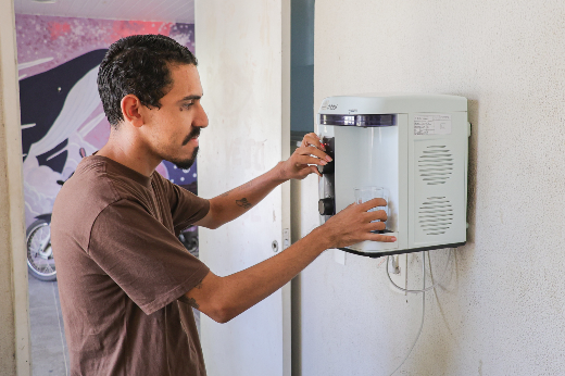 Entre os 65 bens entregues para as Residências Universitárias em Fortaleza estão 44 purificadores de água (Foto: Ribamar Neto/UFC) Imagem: jovem de pele negra com copo de vidro na mão retira água de um purificador de água afixado na parede