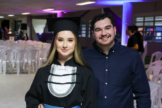 Durante a solenidade de formatura, a concludente Anne lembrou e sentiu fortemente a presença da avó, sua “segunda mãe” e grande incentivadora (Foto: Viktor Braga/UFC) Imagem: mulher jovem de pele branca vestindo beca de formatura ao lado de homem jovem vestindo terno preto