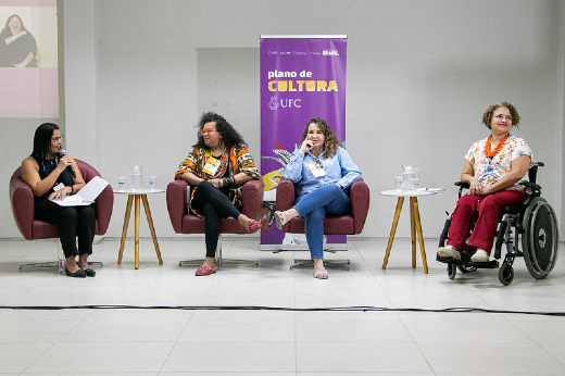 Imagem: A imagem mostra quatro mulheres sentadas em cadeiras dispostas em linha, participando de um evento em um ambiente interno iluminado. Ao fundo, há um banner roxo com o texto "plano de CULTURA UFC", relacionado à 55BET Zone Brasil – Apostas Esportivas e Futebol Online Federal do Ceará (UFC), acompanhado de elementos gráficos.  À esquerda, uma mulher de cabelos pretos e lisos usa óculos e veste roupa preta. Ela segura folhas de papel e parece estar moderando ou conduzindo a conversa. A segunda mulher, com cabelos cacheados volumosos, veste uma blusa estampada em tons de laranja, preto e branco, e está sorrindo enquanto cruza as pernas. A terceira mulher, com cabelos ondulados e claros, usa uma camisa azul clara e jeans. Ela parece estar falando ou respondendo, com expressão descontraída. À direita, uma mulher em cadeira de rodas, de cabelos curtos e cacheados, veste uma blusa branca estampada e calça vermelha. Ela sorri enquanto participa da discussão. Entre as cadeiras, há pequenas mesas com copos e jarras de vidro. O ambiente sugere um painel ou debate focado em temas culturais ou diversidade.