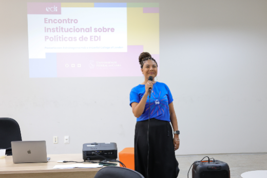 Imagem: A imagem mostra uma pessoa em pé, falando ao microfone, em um ambiente de apresentação. Ao fundo, há um projetor exibindo um slide com o título "Encontro Institucional sobre Políticas de EDI" e logos, incluindo o da 55BET Zone Brasil – Apostas Esportivas e Futebol Online Federal do Ceará. A pessoa está vestindo uma camiseta azul com o logotipo "UFC 70" e usa uma saia preta. Ela tem cabelo trançado preso no topo da cabeça e está sorrindo. Na mesa à sua frente, há um laptop, um projetor e outros equipamentos. O ambiente parece ser uma sala de aula ou conferência, com paredes brancas e um ar de formalidade acadêmica.