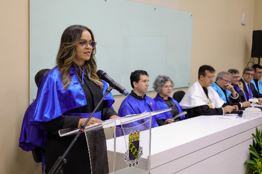 Em seu discurso como oradora docentre, a professora Elisângela da Silva Rodrigues destacou: "Vocês são os pioneiros do nosso legado." (Foto: Viktor Braga/UFC) Imagem: moça de óculos e cabelos castanhos vestindo beca preta e túnica azul discursa em púlpito