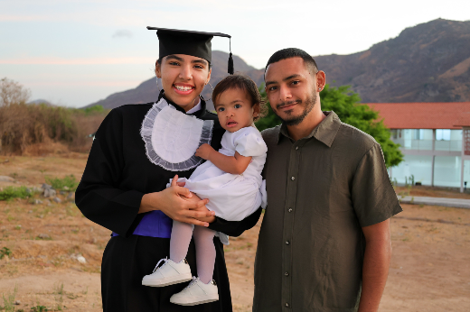 Além do diploma em Análise e Desenvolvimento de Sistemas, Vitoria Nascimento sai da graduação com uma família inteira: do relacionamento com o colega Orácio Cruz nasceu a pequena Agnes (Foto: Viktor Braga/UFC) Imagem: uma mulher em trajes de formatura posa ao lado de sua família. Ela veste uma beca preta com detalhes brancos no peito, uma faixa azul na cintura e um capelo preto. Sorri enquanto segura uma criança de colo, que usa um vestido branco e sapatinhos também brancos. Ao lado delas, um homem com barba e cabelo curto veste uma camisa verde-oliva e também sorri. O fundo da imagem mostra um terreno seco, uma árvore verde, um prédio branco com telhado vermelho e montanhas sob um céu claro.
