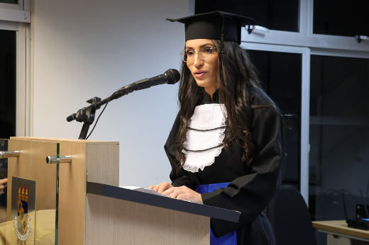 Escolhida oradora discente para a cerimônia de colação, Deigela Lima Rufino ressaltou a importância das diferentes experiências vividas ao longo da graduação (Foto: Viktor Braga/UFC) Imagem: uma mulher está usando uma beca de formatura preta com uma faixa azul e uma gola branca com babados. Ela está de pé atrás de um púlpito, falando em um microfone. A mulher tem cabelos longos e escuros, usa óculos e está olhando para a frente com uma expressão séria e concentrada. Ao fundo, há uma janela de vidro, e o ambiente parece ser uma sala de eventos ou auditório, possivelmente em uma cerimônia de formatura.