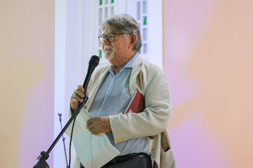 O professor César Barreira falou sobre o processo de construção do livro, elaborado a várias mãos e baseado na Comissão da Verdade UFC e UECE, de 2014 (Foto: Ribamar Neto/UFC) Imagem: um homem idoso de cabelo e cavanhaque grisalhos, paletó cor creme e camisa social azul, fala ao microfone, segurando as folhas com seu discurso nas mãos