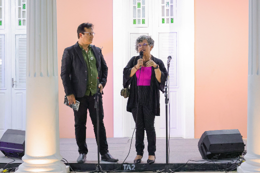 O reitor Custódio Almeida e a vice-reitora Diana Azevedo iniciaram as falas do evento destacando a importância do movimento estudantil e o fato de a Reitoria estar de portas abertas para a comunidade (Foto: Ribamar Neto/UFC) Imagem: duas pessoas em um palco. À esquerda, um homem de óculos usa um terno preto com uma camisa verde e segura um livro ou caderno enquanto observa a pessoa ao lado. À direita, uma mulher de cabelo encaracolado e grisalho, vestindo uma blusa rosa e um casaco preto, fala ao microfone. Eles estão posicionados entre duas colunas brancas, com portas brancas e detalhes em vidro colorido ao fundo, em frente a uma parede rosa. O ambiente sugere um evento formal ou cultural.