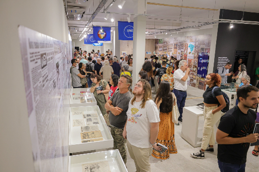 Dezenas de pessoas conferiram a exposição na abertura, e mesmo pessoas que acompanharam e contribuíram para a elaboração do conteúdo se disseram surpresas com o resultado final (Foto: Ribamar Neto/UFC) Imagem: espaço de exposição interna, com várias pessoas caminhando e observando objetos expostos em vitrines de vidro. Ao fundo, há bandeiras azuis com o logotipo da UNE (União Nacional dos Estudantes). O ambiente parece ser iluminado com luz branca, e as paredes estão decoradas com painéis exibindo imagens e textos históricos. O público é diverso, com pessoas de várias idades, algumas interagindo entre si, enquanto outras estão concentradas nos itens expostos. O local transmite uma atmosfera de interesse cultural e histórico.