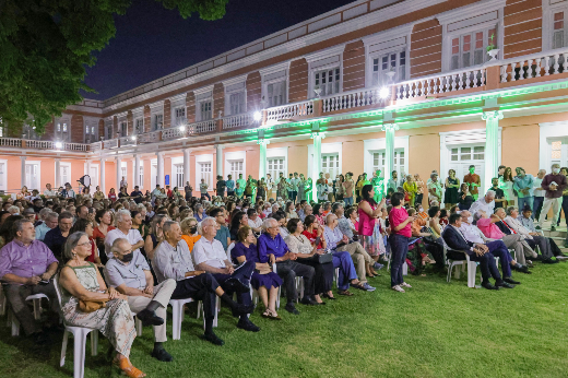 O público lotou os Jardins da Reitoria para ouvir participantes do evento, que falaram sobre democracia, reparação, militância e outros temas relacionados à noite (Foto: Ribamar Neto/UFC) Imagem: evento ao ar livre durante a noite, realizado em frente a um grande prédio histórico de fachada rosa e branca com arquitetura colonial, iluminado por refletores verdes. Um grande público está sentado em cadeiras brancas de plástico, organizado em fileiras sobre o gramado. Entre os participantes, há pessoas de diferentes idades, com algumas em pé e outras conversando. O ambiente é iluminado e festivo, transmitindo a sensação de uma celebração ou encontro cultural.
