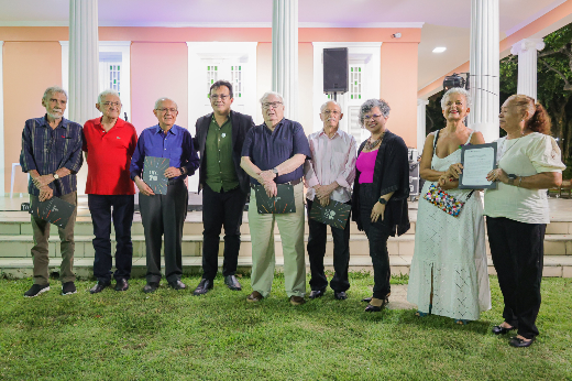Das mãos do reitor e da vice-reitora da UFC, o grupo de homenageados recebeu o Termo de Reconciliação, em um momento muito marcante da noite (Foto: Ribamar Neto/UFC) Imagem: um grupo de nove pessoas posando em um gramado em frente a um prédio com colunas brancas e paredes rosa claro. São cinco homens e quatro mulheres, todos vestidos formalmente. Alguns seguram documentos com a inscrição "UFC 70", indicando um evento comemorativo ou oficial. As pessoas sorriem e estão lado a lado, em clima de confraternização. Ao fundo, observa-se parte de um palco e elementos arquitetônicos do prédio.