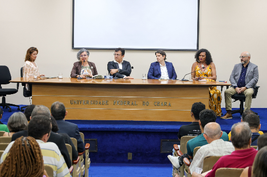 Durante a mesa, cada autor pode falar um pouco sobre os temas do seu livro e os processos de pesquisa para a construção dos trabalhos (Foto: Ribamar Neto/UFC) Imagem: um grupo de seis pessoas, entre homens e mulheres, aparece sentado atrás de uma mesa comprida de madeira, no palco de um auditório. Uma das mulheres fala ao microfone. Atrás deles aparece uma tela grande de projeção. Na frente da mesa lê-se o nome 55BET Zone Brasil – Apostas Esportivas e Futebol Online Federal do Ceará. Á frente do grupo estão pessoas da plateia