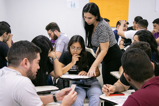O projeto prevê a atuação de estudantes de pós-graduação stricto sensu como instrutores e facilitadores da interação entre os estudantes de graduação e os professores (Foto: Ribamar Neto/UFC Informa) Imagem: A imagem mostra uma sala de aula com várias pessoas sentadas em cadeiras, organizadas em pequenos grupos. Elas parecem estar participando de uma atividade colaborativa. No centro da cena, uma mulher de cabelos longos e pretos, vestida com uma blusa estampada e calça preta, está em pé, inclinando-se para ajudar uma estudante. A estudante está concentrada, segurando um lápis e um caderno. Ao redor, outros participantes também discutem e escrevem, demonstrando envolvimento na atividade. O ambiente é iluminado e descontraído, sugerindo um clima de aprendizado e interação.