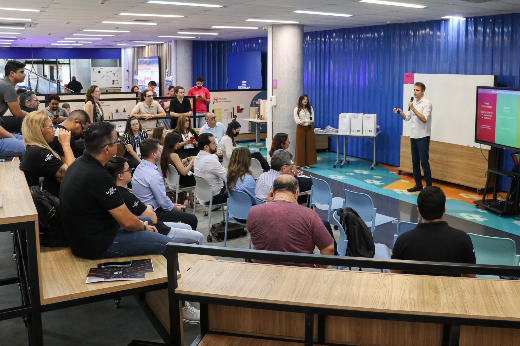 IV Mostra de Startups, realizada em dezembro de 2024 pelo Sebrae Ceará, com apoio da UFC: evento foi uma das ações articuladas pela Prointer após a alteração em seu escopo de atribuições (Foto: Ribamar Neto/UFC) Imagem: um jovem apresentador, vestindo camisa branca e calça jeans, está de pé na frente de uma plateia, segurando um microfone e um controle remoto. Ele gesticula enquanto apresenta slides exibidos em uma tela grande ao seu lado. A plateia, composta por homens e mulheres de diferentes idades e estilos, está sentada em arquibancadas e cadeiras, prestando atenção. O espaço tem um design moderno, com paredes azuis, iluminação clara e um ambiente colaborativo. Ao fundo, há uma mulher de pé perto de algumas impressoras e um grupo de pessoas observando a apresentação