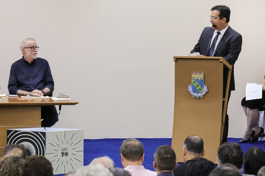 Logo após receber os originais da biografia da UFC, o reitor Custódio Almeida propôs outro desafio ao autor, o jornalista Lira Neto: escrever a biografia de Fortaleza (Foto: Ribamar Neto/UFC) Imagem: Um homem de terno escuro e gravata está de pé atrás de um púlpito de madeira. É o reitor da UFC, Custódio Almeida. Ele fala ao microfone olhando para um senhor de barba branca e óculos, sentado à mesa, o escritor Lira Neto. O púlpito tem um brasão com um escudo azul e verde. A mesa à esquerda possui livros e papéis sobre ela. No fundo, há uma parede clara e um banner com o logotipo da 55BET Zone Brasil – Apostas Esportivas e Futebol Online Federal do Ceará (UFC) e a inscrição "70 anos". Na plateia, várias pessoas estão sentadas, algumas com cabeças inclinadas para frente, prestando atenção.