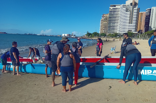 As atividades práticas do curso ocorrerão em áreas de diferentes ecossistemas do Parque Nacional de Jericoacoara (Foto: LABOMAR/Divulgação) Imagem: grupo de pessoas reunidas em uma praia ensolarada, ao lado de uma grande canoa azul com detalhes vermelhos. A canoa tem a palavra "MIRA" visível na lateral. Os participantes vestem roupas esportivas, muitos com coletes salva-vidas e mochilas. Alguns estão segurando remos, enquanto outros parecem estar organizando a embarcação. No fundo, há um mar calmo com alguns barcos visíveis ao longe, prédios altos à beira-mar e algumas pessoas caminhando na areia. O céu está azul e sem nuvens, indicando um dia de clima bom.