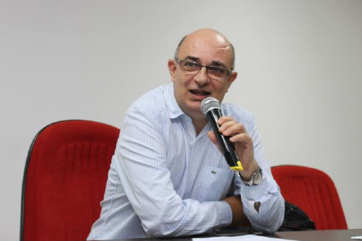 O lançamento de "A pele da história" em Fortaleza será o primeiro no País. Na sequência, Durval Muniz deve passar por outras capitais (Foto: divulgação) Imagem: um homem careca, de ósulos de armação preta e grossa e camisa social quadriculada na cor azul clara fala ao microfone sentado em uma cadeira de escritório com estofado vermelho, atrás de uma mesa. É o professor Durval Muniz