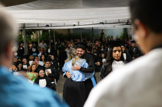 A solenidade lotou o pátio do campus da UFC em Sobral, com participação de amigos, familiares e até pets dos formandos (Foto: Ribamar Neto/UFC) Imagem: Em uma cerimônia de formatura ao ar livre, um formando de beca preta e faixa azul abraça emocionado uma mulher de cabelos loiros, possivelmente uma professora ou familiar. Ambos sorriem enquanto são observados por colegas, professores e convidados sentados ao fundo. O clima é de celebração e conquista