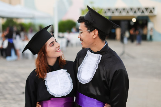 O casal Douglas Freitas do Nascimento e Mayra Beatrisse Freire se conheceu no curso de Música e comemorou juntinho a conquista da graduação (Foto: Ribamar Neto/UFC) Imagem: Um casal de formandos sorri um para o outro com carinho. Ambos vestem becas pretas com golas brancas rendadas e faixas roxas na cintura, além dos capelos (chapéus de formatura). A mulher tem cabelos ruivos e curtos; o homem usa óculos e tem cabelo encaracolado. Eles estão ao ar livre, com outras pessoas e cadeiras ao fundo, em clima de comemoração