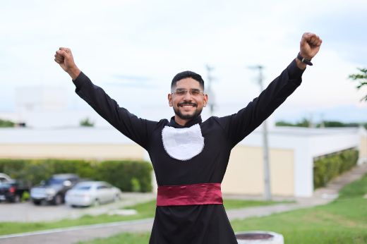 Formado em Odontologia, José Victor Farrapo da Silva comemorou a possibilidade de colar grau com uma cerimônia presencial, ao lado de pessoas queridas: "momento muito aguardado" (Foto: Ribamar Neto/UFC) Imagem: Um homem jovem está em pé ao ar livre, sorrindo com os braços erguidos em sinal de vitória. Ele veste uma beca preta de formatura com uma faixa vermelha na cintura e uma gola branca decorativa. Ao fundo, é possível ver carros estacionados, um prédio claro e vegetação. O céu está claro com algumas nuvens