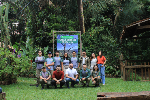 Sob coordenação de Esequiel Mesquita, a equipe é formada por estudantes de pós-graduação, com apoio de guias do Parque Nacional de Ubajara e técnicos do ICMBio (Foto: divulgação) Imagem: Foto de um grupo de 13 pessoas posando ao ar livre em frente a uma placa que diz “Bem-vindos ao Parque Nacional de Ubajara”. O grupo está dividido em duas fileiras: a fileira de trás está em pé e a da frente está agachada. Ao fundo, há vegetação densa, com árvores, plantas tropicais e flores roxas. À direita, vê-se uma estrutura de madeira que parece um pequeno abrigo. O ambiente é verde e natural, transmitindo a sensação de floresta. As pessoas estão sorrindo e vestem roupas casuais, algumas com camisetas do parque.