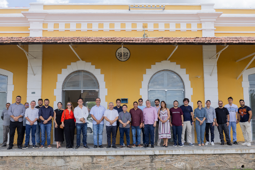 Imagem: Um grupo de 21 pessoas está posicionado em pé, lado a lado, em frente a um prédio histórico de fachada amarela com janelas em arco e detalhes brancos. A construção tem um letreiro circular no alto com o ano "1922". As pessoas estão sorrindo e vestem roupas sociais e casuais em tons variados, como branco, azul, preto, vermelho e bege. O clima parece agradável, com céu parcialmente nublado.
